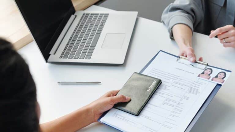 A person hands over a passport and a visa application form on a desk, with a laptop and a pen in the background.