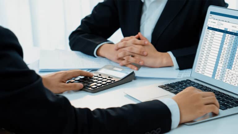 Geschäftsleute analysieren Finanzdaten auf einem Laptop und verwenden einen Taschenrechner zur Berechnung der Mehrwertsteuer.