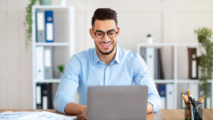 Smiling businessman in glasses working on a laptop at an office desk with financial reports and binders